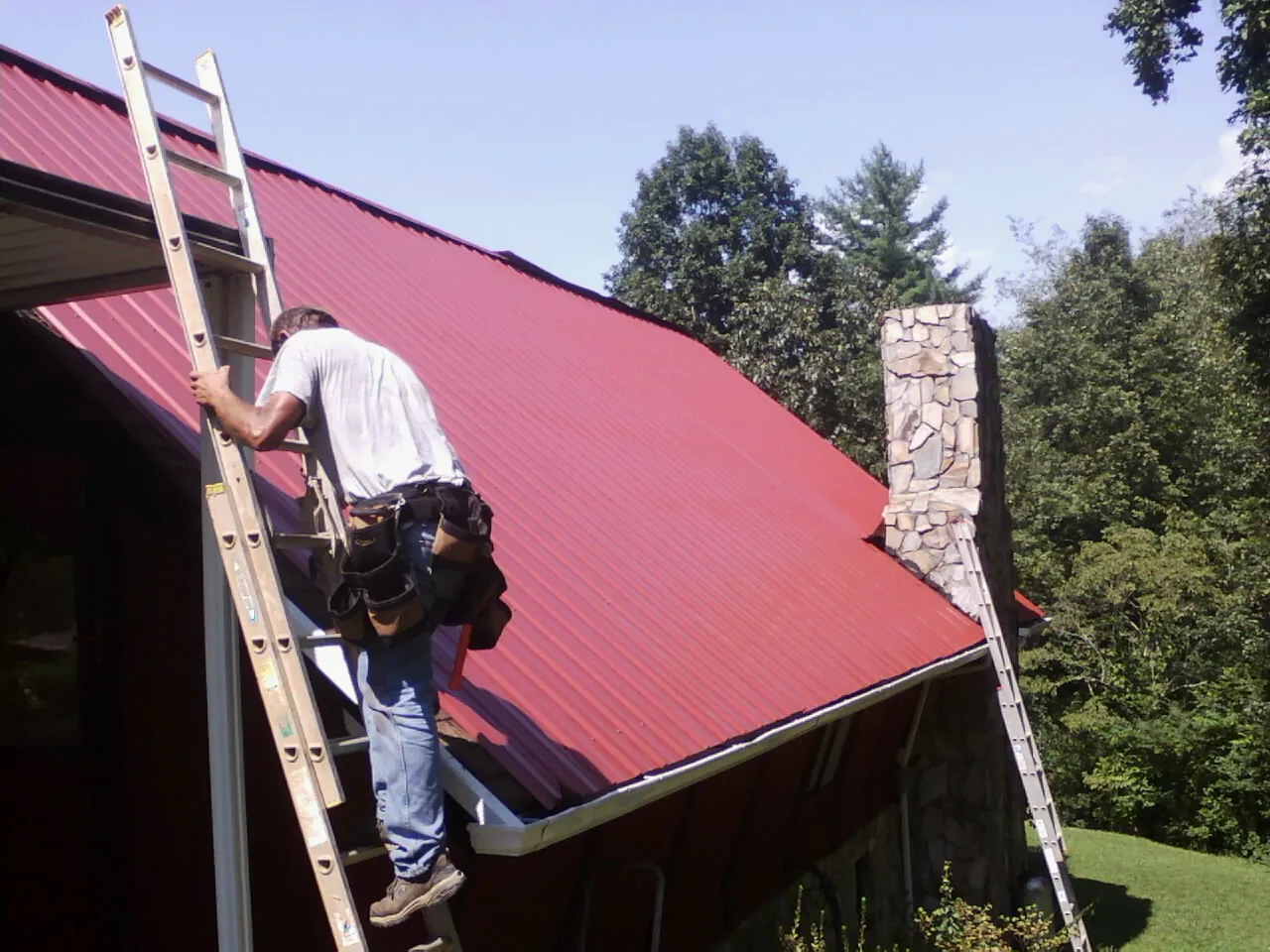 Roofer on a ladder beside a red metal roof and stone chimney — Soffit & Fascia Repair by Roofing Brothers in Orrtanna